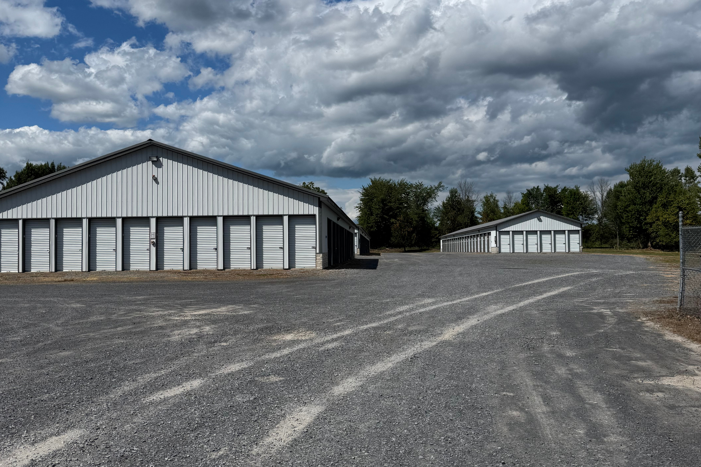 Wide-angle view of several self-storage buildings with white roll-up doors, surrounded by gravel lot and trees in the distance under a cloudy sky.